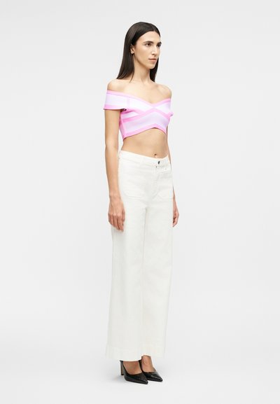 Γυναίκα με μακριά σκουρόχρωμα μαλλιά, ντυμένη με ροζ crop top χωρίς ώμους, λευκό παντελόνι ψηλόμεσο με φαρδιές παντελόνες και μαύρες μυτερές γόβες, στέκεται πλαγιαστά.