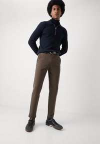 Mørkeblå hettegenser med glidelås, brune bukser og svarte joggesko. Buksene har en glatt tekstur, mens genseren har krage og er slim fit.