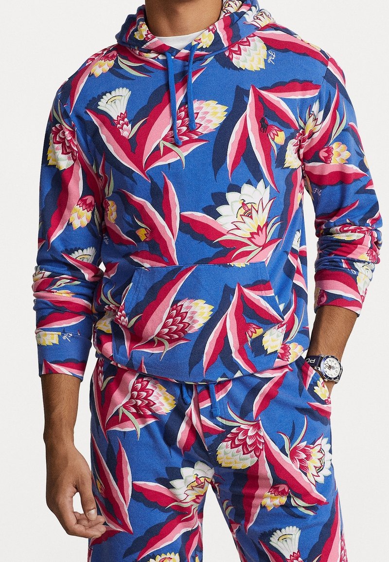 Homme portant un sweat à capuche bleu et un pantalon assorti avec un grand imprimé floral rose, blanc et jaune, mains dans les poches, et montre au poignet gauche.