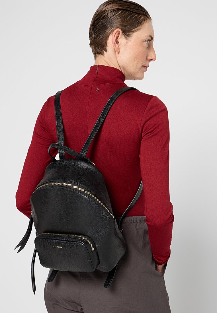 Zaino in pelle nera con forma arrotondata, dettagli in zip dorate e spalline regolabili. Presenta una piccola tasca frontale e una superficie testurizzata.