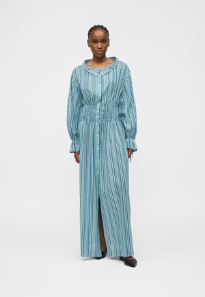 Kobieta stojąca, ubrana w długą sukienkę w pionowe niebiesko-białe paski z guzikami, z marszczonym pasem i rozszerzanymi mankietami, w połączeniu z czarnymi spiczastymi butami.