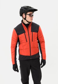Veste isolante rouge et noire avec fermeture éclair à l'avant, deux poches sur la poitrine et manches longues, accompagnée de gants noirs et d'un casque.