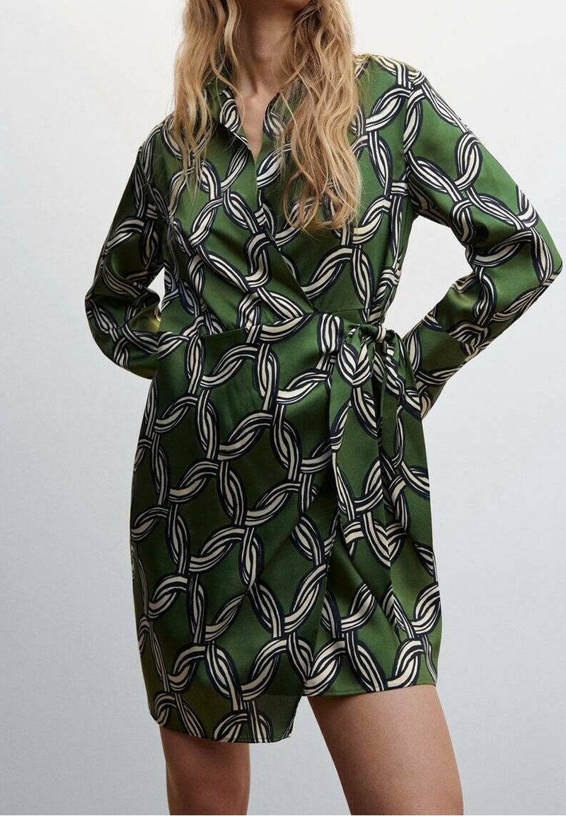 Femme en robe portefeuille verte à manches longues avec un motif chaîne noir et blanc, debout les mains sur les hanches devant un fond uni.