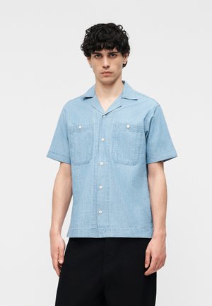 Jeune homme aux cheveux bouclés foncés portant une chemise à manches courtes bleu clair avec deux poches poitrine et un pantalon noir.