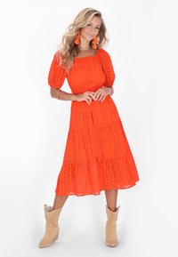 Rochie midi portocalie cu detalii perforate, mâneci bufante, design în straturi și curea din același material. Asortată cu cizme cowboy cu toc de culoare bej și cercei florali.