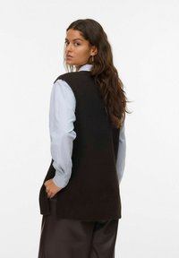 Bruine gebreide mouwloze trui met een ronde halslijn, gedragen over een lichtblauw long-sleeve shirt, met een relaxte pasvorm en zijsplitten.