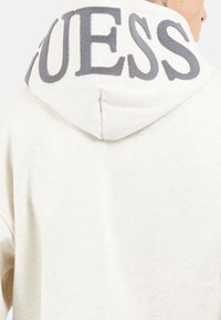 Sudadera color crema con un logo "GUESS" texturizado en gris en la capucha, con una tela suave y un diseño de corte relajado.