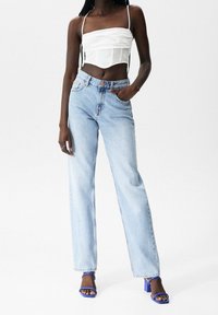 Lichtblauwe high-waisted jeans met wijde pijpen, gecombineerd met een witte cropped top. Model draagt blauwe block-heeled sandalen, waarbij de tenen zichtbaar zijn.