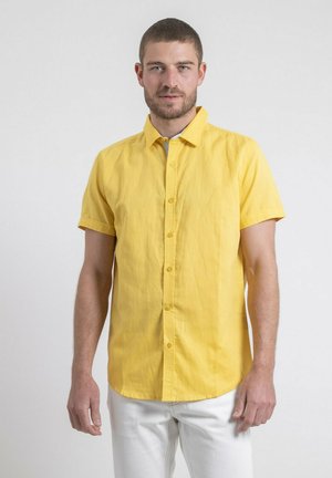 Homme portant une chemise jaune à manches courtes boutonnée et un pantalon blanc, debout devant un fond uni gris clair.