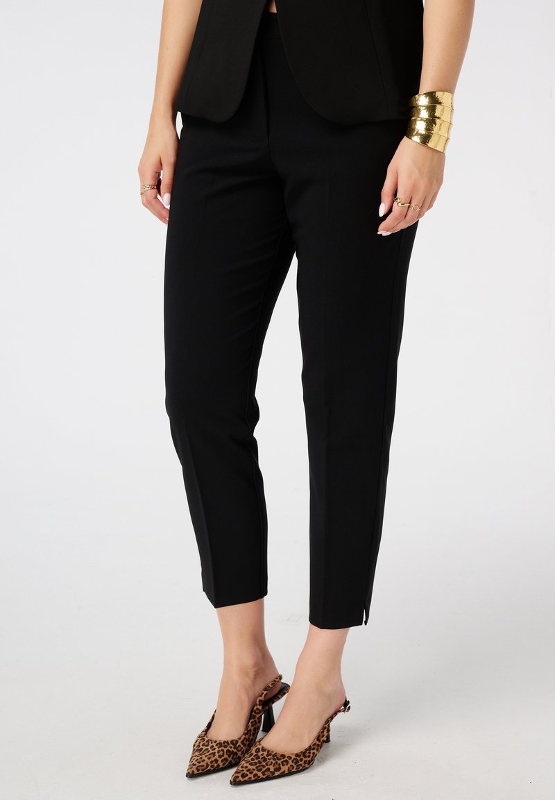Pantalons noirs ajustés avec une coupe slim, présentant une texture lisse et une longueur chevillée. Associés à des talons à imprimé léopard et à des accessoires dorés.