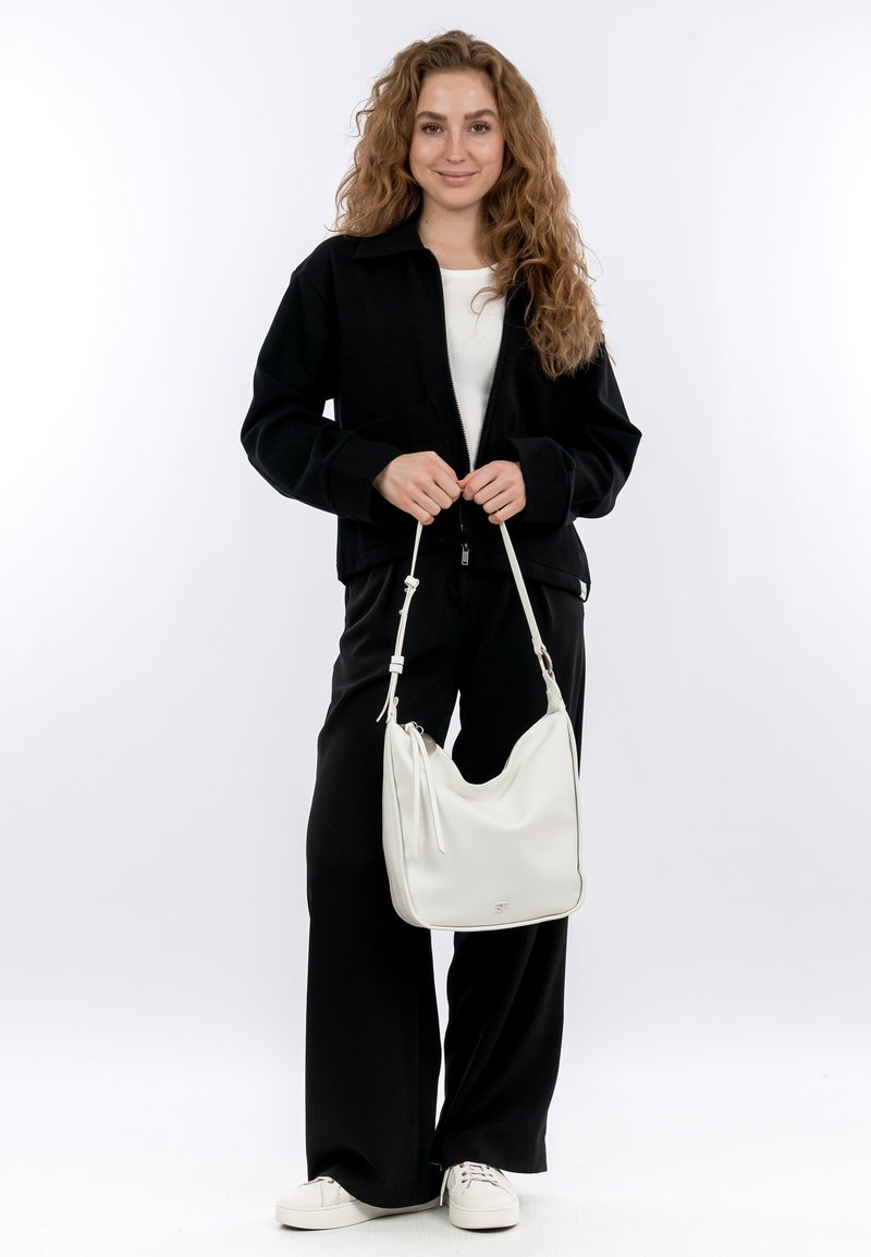 Jacket nera con zip sopra una maglietta bianca, abbinata a pantaloni neri a gamba larga e sneakers bianche. Una borsa a spalla in pelle bianca completa il look.