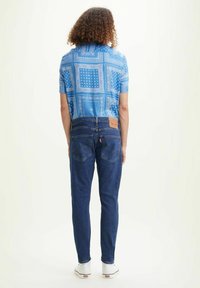 Camisa de manga corta azul con estampado, jeans de mezclilla azules, vista trasera, con etiqueta de marca visible, y zapatillas blancas sobre un fondo claro.