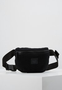 Czarna torba na ramię z polaru z zamkiem na górze, regulowanym paskiem i małą naszywką z logo. Miękka faktura, zaokrąglony kształt.