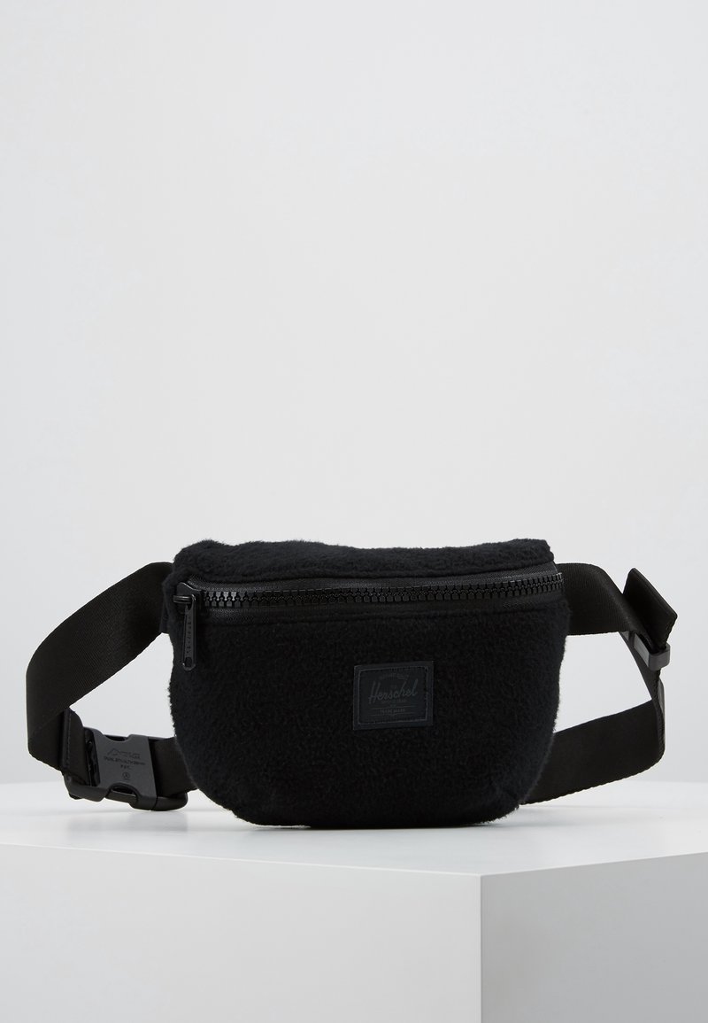 Czarna torba na ramię z polaru z zamkiem na górze, regulowanym paskiem i małą naszywką z logo. Miękka faktura, zaokrąglony kształt.