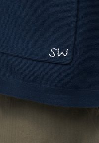 Περδίκι σκούρου μπλε πλεκτού πουλόβερ με λευκά κεντημένα αρχικά "SW" πάνω από ελαιώδη πράσινα υφασμένα παντελόνια.