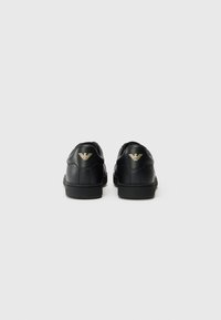 Sorte lærersneakers med en glat tekstur, som har et lavt design, gummisål og et guldlogoemblem ved hælen.