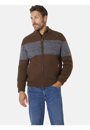 Mann trägt einen braunen Strickpullover mit Reißverschluss und blau gemustertem Streifen über der Brust und blaue Jeans, steht mit einer Hand in der Tasche.