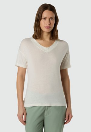 Donna con capelli castani fino alle spalle che indossa una maglietta bianca a maniche corte con scollo a V e pantaloni verde chiaro su uno sfondo semplice.