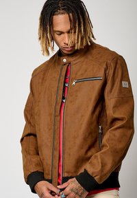 Veste aviateur en cuir marron avec fermeture éclair à l'avant, accents perforés et col montant. Comprend des poches zippées sur les côtés et la poitrine.