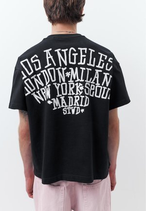 Személy fekete pólót visel, amelyen nagy fehér betűkkel városnevek szerepelnek: Los Angeles, London, Milánó, New York, Szöul, Madrid, egyszerű háttér előtt.