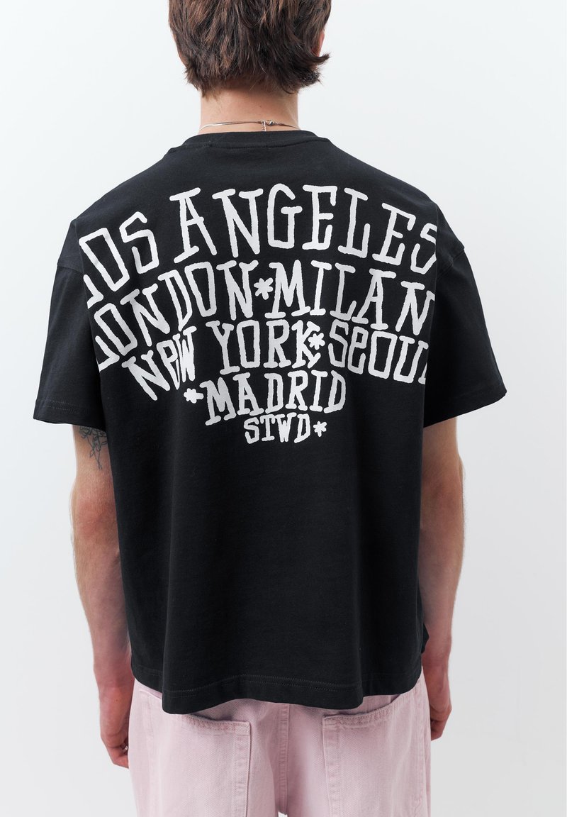 Personne portant un T-shirt noir avec un grand texte blanc listant des villes : Los Angeles, Londres, Milan, New York, Séoul, Madrid, sur un fond uni.