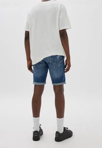 Bílý bavlněný tričko nošené s džínovými šortkami střední praní. Šortky mají ohrnuté manžety a zadní kapsy. Černé tenisky s logem.