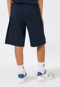 Kind staat, gekleed in marineblauwe shorts, witte kniekousen met logo, en witte sneakers met blauwe accenten en logo.