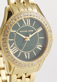 Die goldfarbene Uhr verfügt über ein grünes Zifferblatt mit römischen Ziffern und ist von Kristallen umgeben. Das Armband besteht aus poliertem Metall im Gliederdesign.
