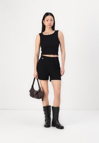 Top negro sin mangas y corto con bordes en cascada, pantalones cortos negros ajustados, botas negras hasta la rodilla y un pequeño bolso marrón de hombro.