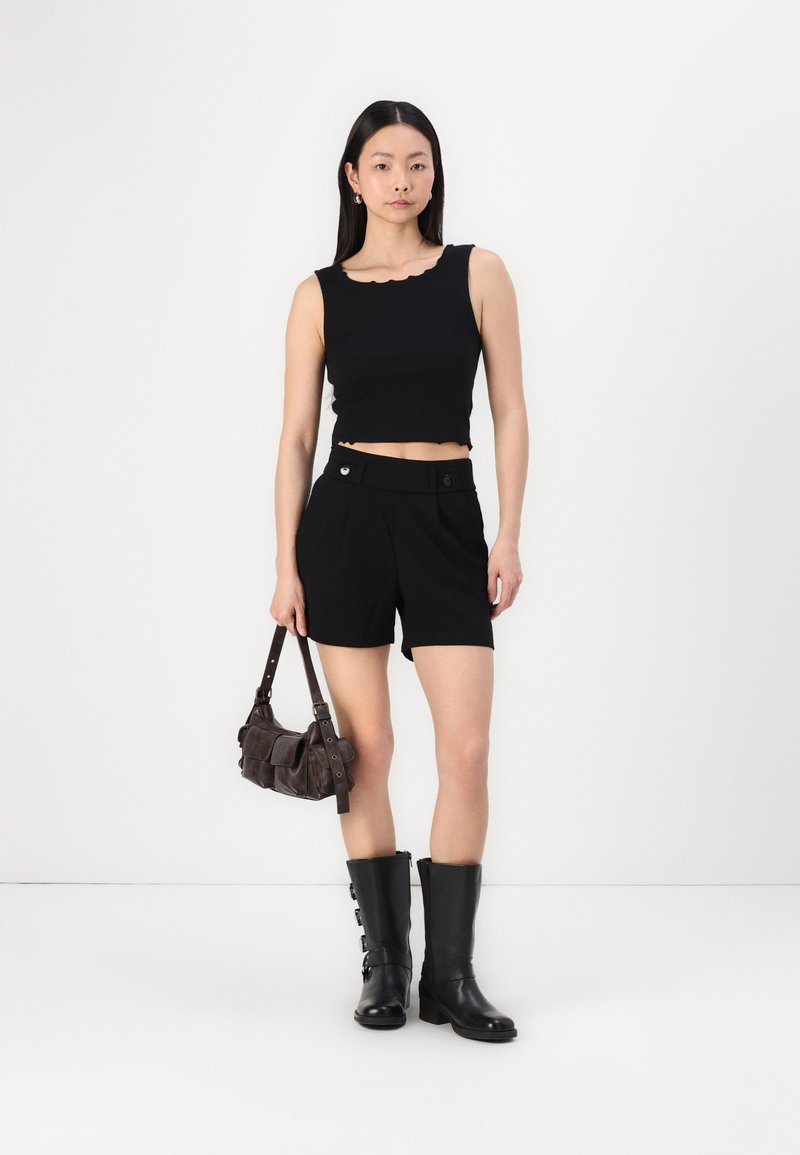 Top negro sin mangas y corto con bordes en cascada, pantalones cortos negros ajustados, botas negras hasta la rodilla y un pequeño bolso marrón de hombro.