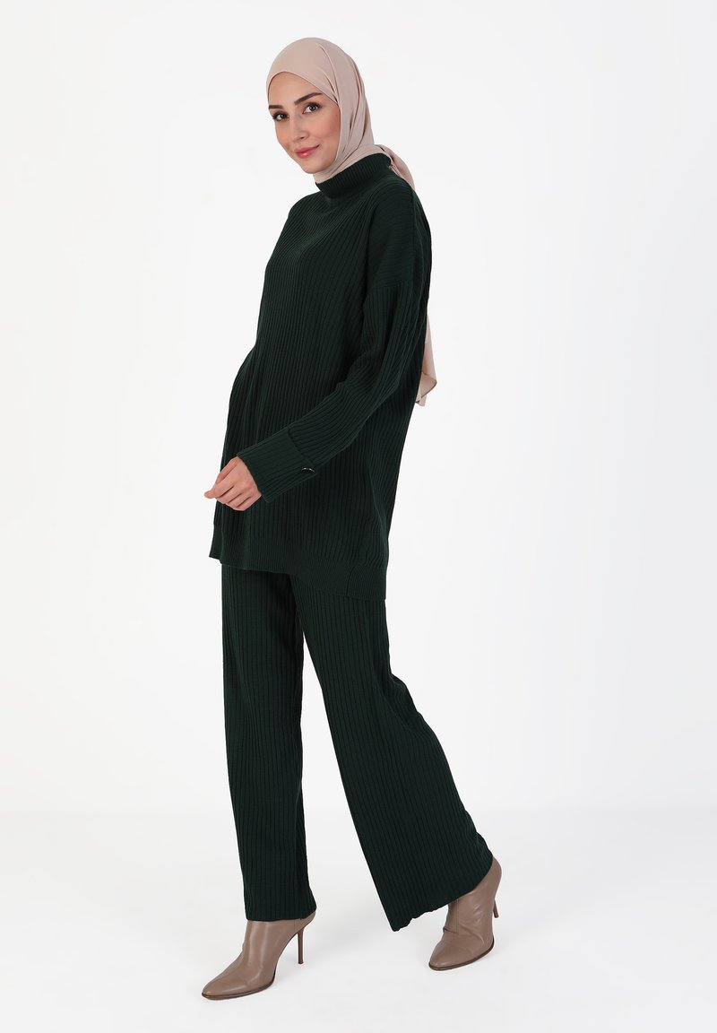 Completo in maglia a coste verde scuro, composto da un top a collo alto e fit morbido, e pantaloni a gamba larga. Un hijab di colore chiaro completa l'outfit.