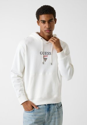 Jeune homme ajustant la capuche d'un sweat à capuche blanc de la marque Guess Jeans, portant un jean bleu clair, debout devant un fond clair uni.