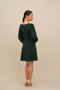 Kvinne med mørkt hår i en lav hestehale iført en mørkegrønn knelang kjole med gjennomsiktige puffermede ermer og nakne høyhælte sandaler, vendt mot høyre.