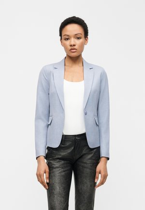 Frau mit kurzen lockigen Haaren, die einen hellblauen Blazer, ein weißes geripptes Top und schwarze strukturierte Hosen trägt, steht vor einem schlichten weißen Hintergrund.