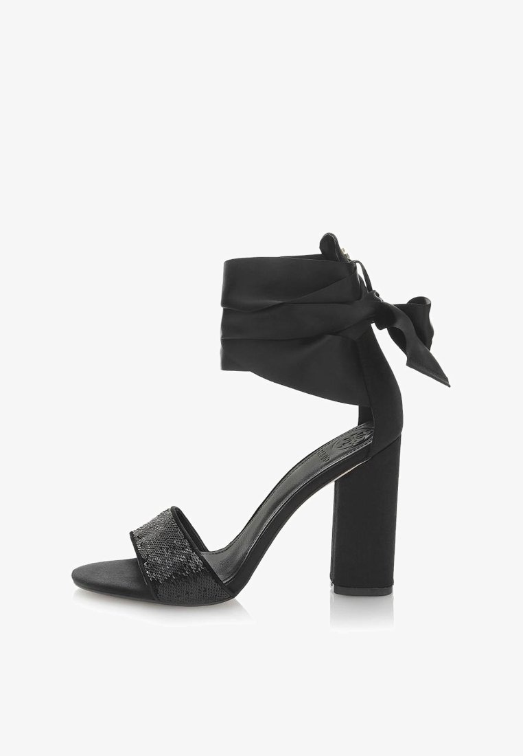 Czarny but na wysokim obcasie, z blokowym obcasem, materiałowym zapięciem w okolicy kostki i strapem pokrytym cekinami. Posiada gładką fakturę i odkryty czubek.