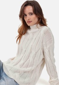 Grober, cremefarbener Strickpullover mit hohem Kragen, der strukturierte Muster und eine lockere Passform aufweist, kombiniert mit hellblauem Denim.