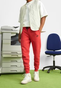 Röda sweatshirtbyxor med sidfickor och dragsko i midjan, kombinerat med en lätt vadderad väst och vita sneakers, mot en kontorsbakgrund.