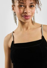 Mlađa žena s kovrčavom kosom koja nosi duge kristalne naušnice i crnu baršunastu haljinu s tankim perlicama, blago se smiješi.