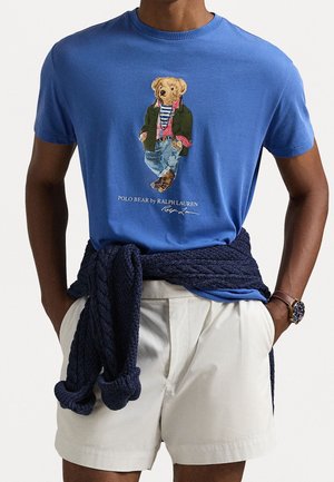 Personne portant un t-shirt bleu avec un motif d’ours en peluche, un short blanc et un pull bleu marine noué autour de la taille.