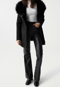 Mujer vestida con abrigo negro forrado de piel, blusa blanca ajustada, pantalones de cuero negros y botas de tacón negras, de pie frente a un fondo liso.