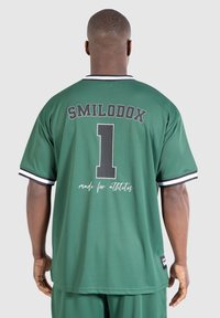 Muž v zeleném sportovním dresu a šortkách, pohled zezadu, s nápisem "SMILODOX" a velkým číslem "1" vytištěným na drese.