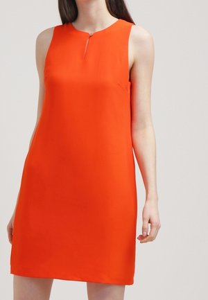 Frau trägt ein ärmelloses, leuchtend oranges Kleid mit kleinem Reißverschlussdetail vorne am Ausschnitt und steht vor einem einfarbigen Hintergrund.