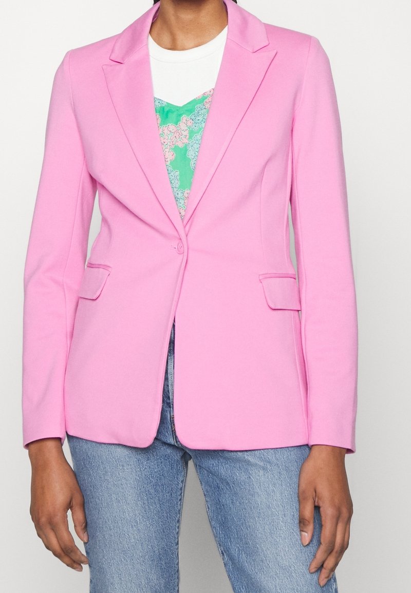 Personne portant un blazer rose cintré sur un haut vert à fleurs et un jean bleu clair, debout avec les bras détendus le long du corps.