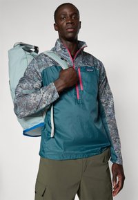 Teal halv-zip jakke med mønstrede grå ermer, som har en rosa glidelås. Modellen bærer en lyseblå ryggsekk.