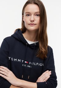Junge Frau mit langen braunen Haaren trägt einen dunkelblauen Tommy Hilfiger Hoodie und steht mit verschränkten Armen vor einem schlichten weißen Hintergrund.