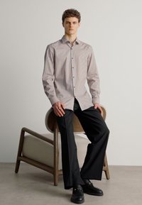 Camicia a maniche lunghe con fantasia leggera, abbinata a pantaloni neri a gamba larga. Modello seduto su una sedia di legno.