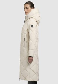 Lungo cappotto beige trapuntato con cappuccio, caratterizzato da cuciture diagonali, tasche laterali e una patch nera con logo sulla manica sinistra. Chiusura a zip.