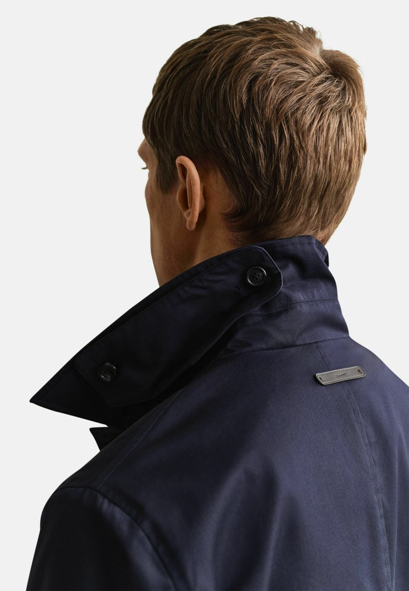 Ein Mann, von hinten gesehen, trägt einen marineblauen Mantel mit hochgeschlossenem Kragen und hat kurzes, hellbraunes Haar.