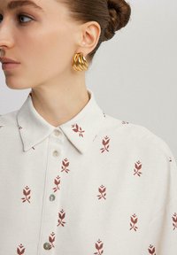 Camicia bianca con stampa floreale rossa, dotata di chiusura con bottoni e tessuto testurizzato. Orecchini a cerchio dorati completano l'outfit.
