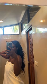 Vit handduk hängd runt axlarna, långt svart hår, smartphone hålls i handen. Ljust badrum med beige kakel och trädörr.